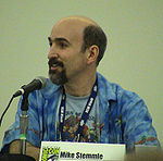 A Caucasian bald man with a black beard looks to the left while speaking into a microphone. A plaque with the Comic-Con logo on the table reads "Mike Stemmle".