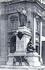 A stone statue of a robed man with hand raised, atop a plinth outside a stone building.