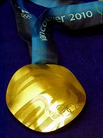 A shiny golden medal is pictured in front of a blue background. The medal has a blue and black ribbon with white writing on it.