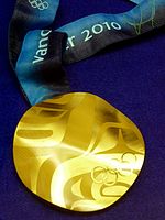 A shiny golden medal is pictured in front of a blue background. The medal has a blue and black ribbon with white writing on it.