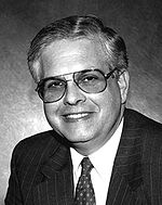 The head and shoulders of a smiling clean-shaven man with graying hair, wearing tinted aviator-style glasses, and a tie and suit jacket with pin-stripes