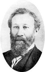 The head of a man with a long curly full beard, his hair parted on his left