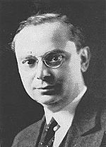 The head and shoulders of a clean-shaven man with hair parted on his left, wearing round, wire-rimmed glasses and a suit and tie