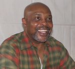 Bald, bearded, smiling African American man in his sixties, wearing a colorful plaid shirt.
