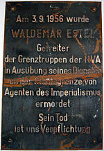Damaged metal plaque reading "Am 3.9.1956 wurde WALDEMAR ESTEL Getreiter der Grenztruppen der NVA in Ausübung seine Dienstes an der Staatsgrenze von Agenten des Imperialismus ermordet. Sein Tod ist uns Verpflichtung."