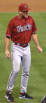 A man wearing gray baseball pants, a red jersey with "D-Backs" written across the chest in black letters, and a red cap with a black "D" on it stands on a baseball field