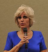 A photograph of a blond-haired, brown-eyed, middle-aged woman in a blue blouse speaking into a microphone.