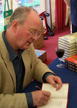 A balding man wearing rimless glasses is sitting at a table autographing books. He is wearing a sports coat. A glass of wine and a stack of books are on the table.