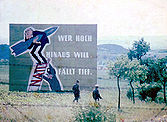 Two armed East German soldiers, seen through a barbed-wire fence, walking from right to left through a grassy hilly landscape towards a clump of young trees. Behind them is a very large propaganda sign showing a caricature of West German Chancellor Konrad Adenauer clutching a missile while standing on a ladder being propped up by a military officer. The rungs of the ladder are made from the acronym "NATO". The sign is captioned: "Wer hoch hinaus will, fällt tief!"