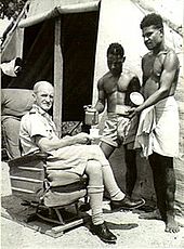 Caucasian man in light-coloured military uniform seated outside tent and being served tea by two indigenous men in light-coloured shorts and loincloth