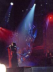 A brunette in a black-and-red kimono moving to the right of a stage with red-lit backgrounds and a metallic tree like structure in the middle. A single source of light falls on her.