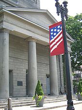 A tall, grey brick building with four columns before the entrance. In the foreground, a black lightpost is seen with a banner featuring a version of the flag of the United States.