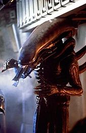 A tall human-like alien creature stands inside the cargo hold of a spacecraft. It is all black in color, has a long, horizontally configured phallic-shaped head with its long rigid tongue extended, long arms with six digits on each hand, and long tubes protruding from its back.