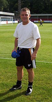 A blond-haired man in his 40s, wearing a white T-shirt and black shorts, stands near the edge of a football pitch.