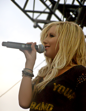 A young blond female singer on a stage sings into a microphone, holding it with her right hand. She is using a black blouse with the words "Your" and "Band" printed on it in a yellow color and few bracelets on her right hand.