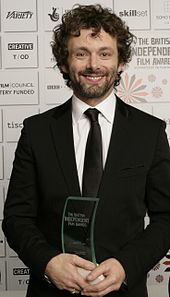 Michael Sheen, a caucasian male in his late-30s with dark shaggy curly hair. He wears a black suit and white shirt with a black tie. He smiles and holds a glass award with both his hands.
