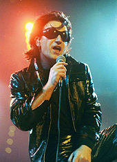 Bono with black hair, black sunglasses, and a black leather attire speaks into a microphone.