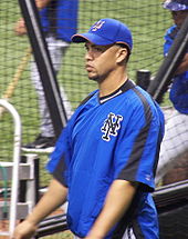 A man in his early thirties wearing a blue warm-up jacket and blue baseball cap rides on a motorized cart. His jacket has black stripes on the arms and "NY" in black on the breast.