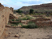 A partly overcast sky and subdued sunlight over a roughly six-foot tall wall of dusky tan sandstone bricks which vary somewhat in size. The wall runs diagonally from the immediate foreground at left towards the right, running perhaps several dozen feet to the near middle distance. A few feet to the right, in the middle foreground, a low ring of similar blocks delimits a circular pit sunk into the ground. The remains of several other ruinous low walls, perhaps one to three high at most, are arrayed in parallel; they align left to right from the high diagonal wall. Perhaps a mile distant to the center and right, a canyon wall slopes gradually level to meet the valley floor on which the walls sit.