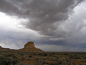 Dark, rolling storm clouds lower over a desert landscape; a butte stands in the near distance, left of center.