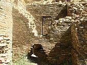 Inside daytime view of a ruined and ceiling-less rectangular room. Tawny-beige stacked sandstone bricks compose walls rising from brush-covered ground. The several walls visible in the image are up to perhaps a dozen feet in height. In the wall immediately at center, a triangle-shaped entrance several feet high leads to an adjacent chamber behind. The upper part of the same wall, shaped like an inverted-triangle, has fallen away or otherwise been removed, revealing a rectangular doorway leading to yet another concealed room. At left and right are two similar walls perpendicular to the one at middle.