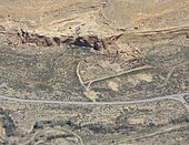 Daytime birds-eye view of a desert with semicircular complex of ruined walls abutting a mesa, which covers the top third of the photo. The straight side of the complex faces a metaled road delimiting the bottom third of the image. The complex itself comprises many smaller enclosed circular and rectangular spaces. These "rooms" no longer have ceilings.