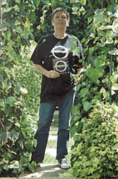 A middle-aged woman stands on a path amidst green vegetation, under bright sunlight.  She wears a black T-shirt with a white logo, and holds a small piece of black card which carries the same logo.  She has blue jeans, and white shoes.  Her hair is cut short.  She is looking slightly upward, over the head of the photographer.