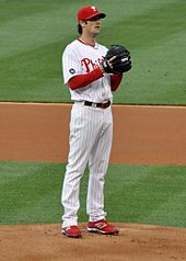 A left-handed man wearing a white baseball uniform and red baseball cap prepares to throw a baseball from a pitcher's mound.