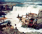 Men sit on scattered wooden platforms surrounding a waterfall that is about 25 feet (7 meters) high and 250 feet (70 meters) wide. The men hold long poles attached to nets that are in the water.