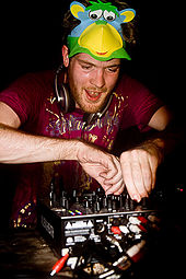 A white man with red hair and a slight beard manipulating the controls of a piece of music mixing equipment.  He has headphones around his neck and a mask resembling a multi-coloured monkey pushed up onto the top of his head.