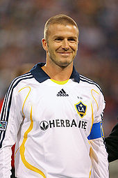 A photograph of a smiling man with a shaven head. He is wearing a white shirt with yellow trim and a navy blue collar, and a light blue armband.