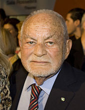 A squarish faced man in a suit and tie: he is balding with closely cropped white hair, and sports a stubble of white beard.