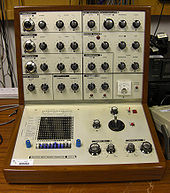 A portable L-shaped brown wooden case with a silver metal fascia filled with buttons and controls is positioned on a wooden work surface.  The controls for the device are mostly rotary, and denoted with lettering and numbering.  The lower part of the box contains a small matrix of holes and a joystick. Other pieces of electrical equipment are visible behind the device.