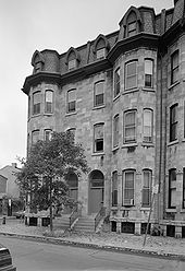 End section of a four-story stone terraced building. Two adjacent doors are at center, each with a short flight of stairs leading up from the street to the first level. Either side of the doors, at left and right, the building has two rounded tower-like sections, each with nine large sash windows (three on each level). Four more large windows are at center, above the doors. The uppermost (attic) level has four smaller windows protruding from the roof with architectural feature. At street level, grilled skylights illuminate basement rooms.