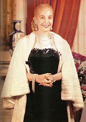 A picture of a Evita, former first lady of Argentina. Her hair is drawn into a tight bun at the back. She is wearing a black, low-cut dress. Around her neck is a number of chains. The lady's hands are folded in her front and she has a white fur shawl around her.