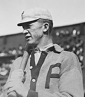 A man in a baseball cap and V-neck buttoned sweater looks to the left of the image. His sweater and his hat both display the letter "P" in block type, and he holds a baseball bat over his shoulder with both hands.