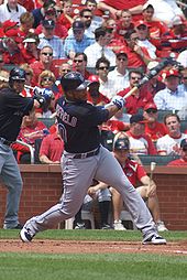 A dark-skinned man in a black baseball jersey and gray pants takes a right handed baseball swing with a crowd in the background, several people wearing red.