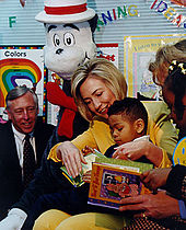 Same woman reads a book in a classroom to an African American boy in her lap, as an African American girl and two adults look on
