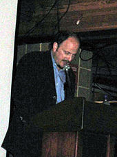 A balding and bearded man stands bowed over a podium.