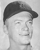 A young man in his mid-twenties smiling and looking to the right of the image; he is wearing a dark baseball cap on his head with an Old English "D" on the front