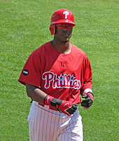 A dark-skinned man in a red baseball jersey and red left-handed batting helmet walks on a baseball field; he appears to be in his mid-twenties. His jersey reads "Phillies" in white and red script, with two blue starts dotting the "i"s.