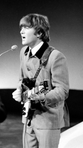Monochrome image of John Lennon playing guitar and speaking into a microphone while wearing a grey suit.