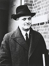 Half-length outdoor portrait of man in dark suit, overcoat and hat
