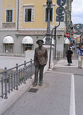 Bronze statue of Joyce standing on a sidewalk, next to a railing. Behind the statue is a street scene with pedestrians and stores.