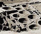 Nine large round pits are seen from above, in washed-out daylight. Eight of the pits descend from a common stone platform; the ninth sits alone on a somewhat higher stone surface. The depressions run diagonally from bottom right to middle top. At bottom left are seen perhaps eight smaller, regularly sized rectangular "rooms" enclosed by ruined walls; to their left are larger ruined enclosures. Bordering the rings at top and right are various smaller rooms and walls which appear less ruined.