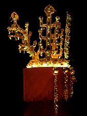A shiny gold crown resembling trees, exhibited in a very dark room. It has beads of rock crystal and translucent and curved green jade ornaments