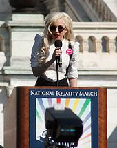 A blond woman wearing a white shirt and black glasses speaking on a lectern carrying a 'National Equality March' poster. Behind her is a white stone balustrade of a building.