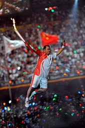 An Asian man in red and white athletic shirt and shorts, and wearing sneakers, is suspended by wires in the air while holding a lit torch. In the background, a large crowd in a stadium can be seen, as well as two blurred flags.