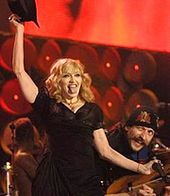 A blond woman in a black dress, holding a black hat atop her head with her riht hand, and a microphon in her left. She is pointing her tongue towards the camera. Beside her the smiling face of a man is visible.