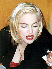 The image of a young blond woman. She is wearing a black coat. Her hair is short, straight and parted from the left to the right. She has bright red lips and appears to be speaking to someone on her left while looking down.
